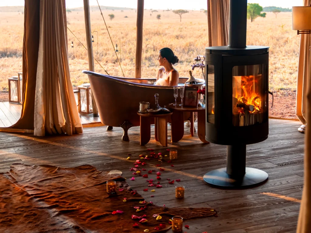 A woman in a copper bathtub overlooks the Serengeti, with a fireplace and rose petals enhancing the luxurious ambiance.