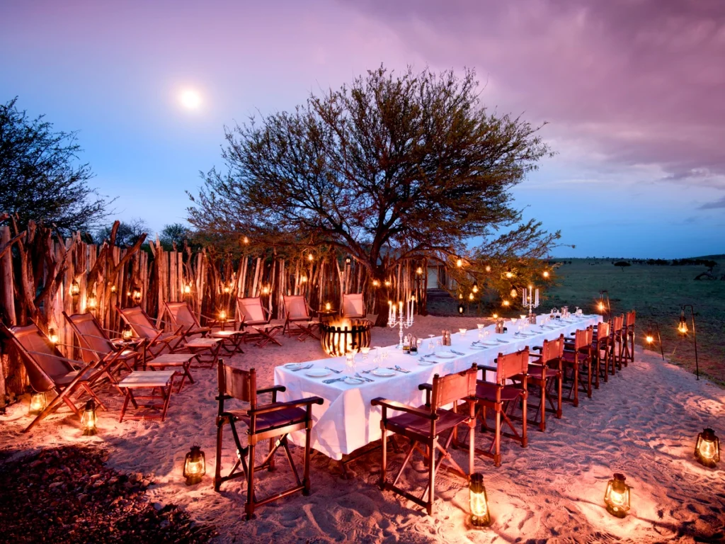 A candlelit dinner setup in the Serengeti wilderness, featuring elegant table decor, soft lantern lighting, and a starry night sky above.