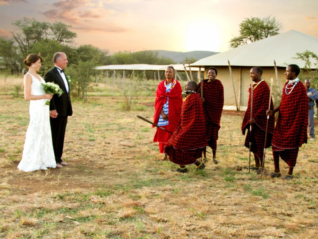 A couple in wedding attire stands before Maasai warriors in traditional red attire, set against a Serengeti sunset with luxury tents in the background.