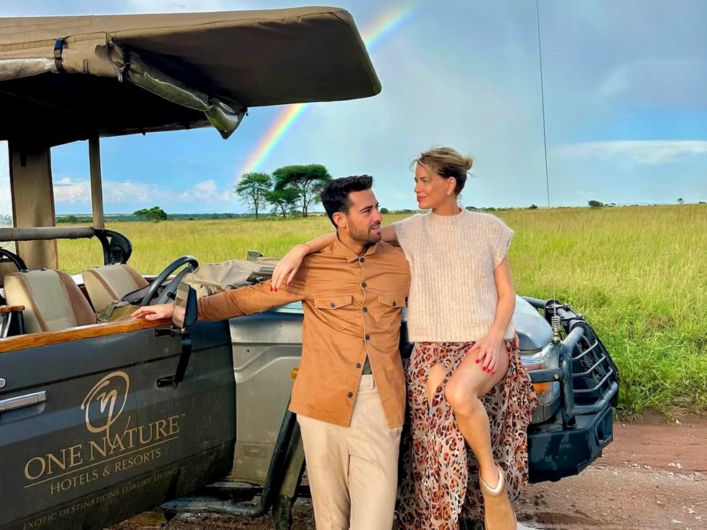 Beautiful couple standing together in Serengeti National Park, facing each other near a One Nature Hotels safari jeep with a rainbow in the background.