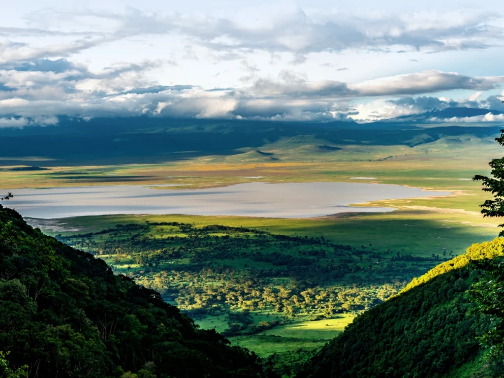 A panoramic view of the Ngorongoro Crater featuring a sparkling lake surrounded by lush greenery under a cloudy sky.